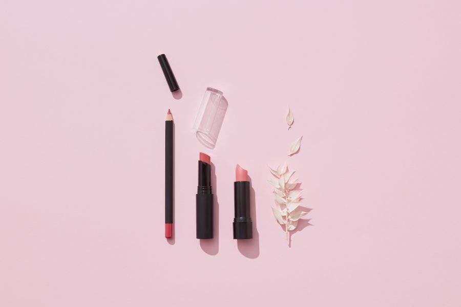 Lipsticks and a lip pencil arranged with a dried leaf on a pink background.